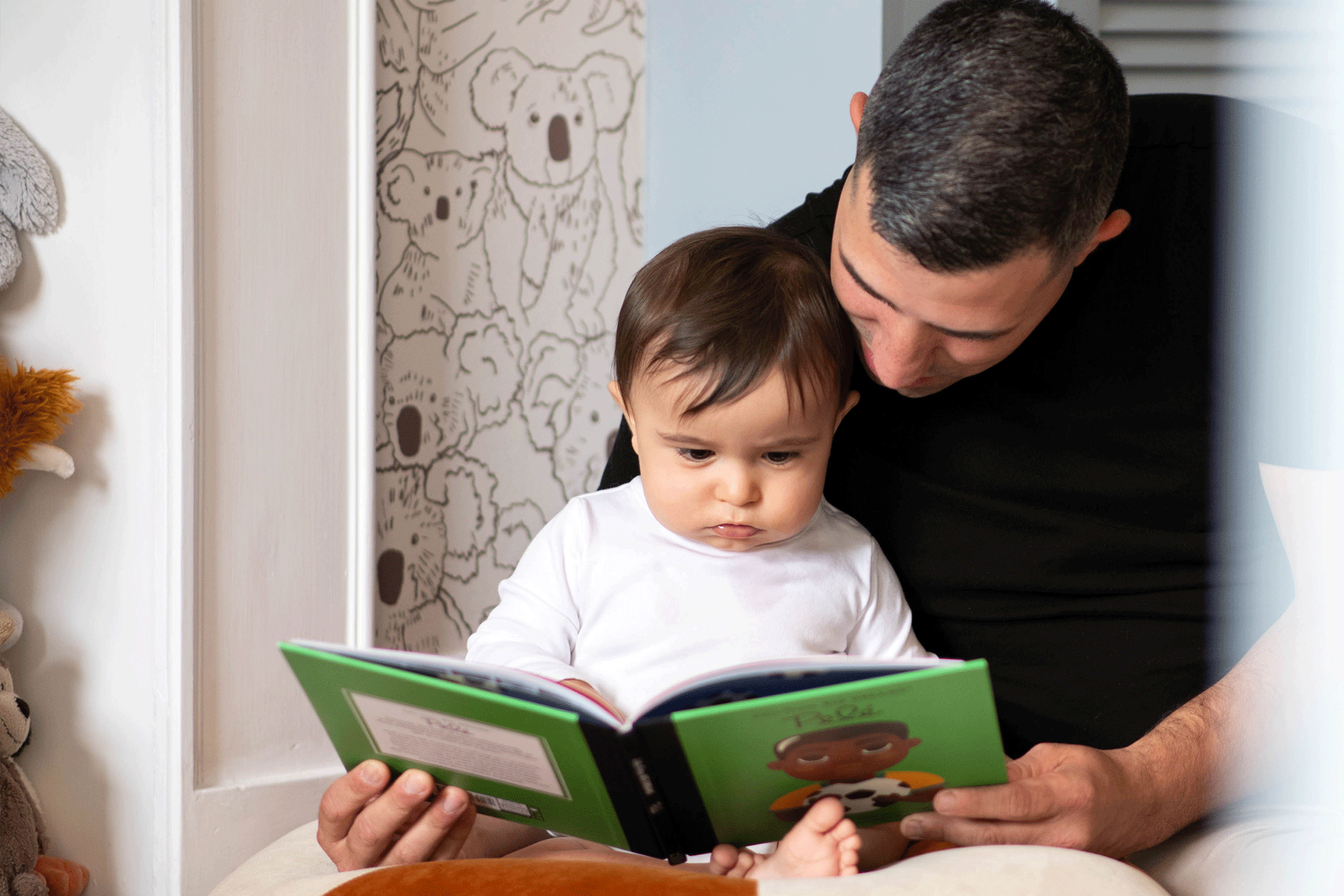 A father sitting with his young toddler on his lap, both looking intently down at an open children's picture book with a green cover. The toddler wears a white long-sleeved shirt, while the father wears a black t-shirt. In the background, a wall features black-and-white illustrated koala wallpaper.