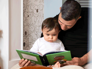 A father sitting with his young toddler on his lap, both looking intently down at an open children's picture book with a green cover. The toddler wears a white long-sleeved shirt, while the father wears a black t-shirt. In the background, a wall features black-and-white illustrated koala wallpaper.