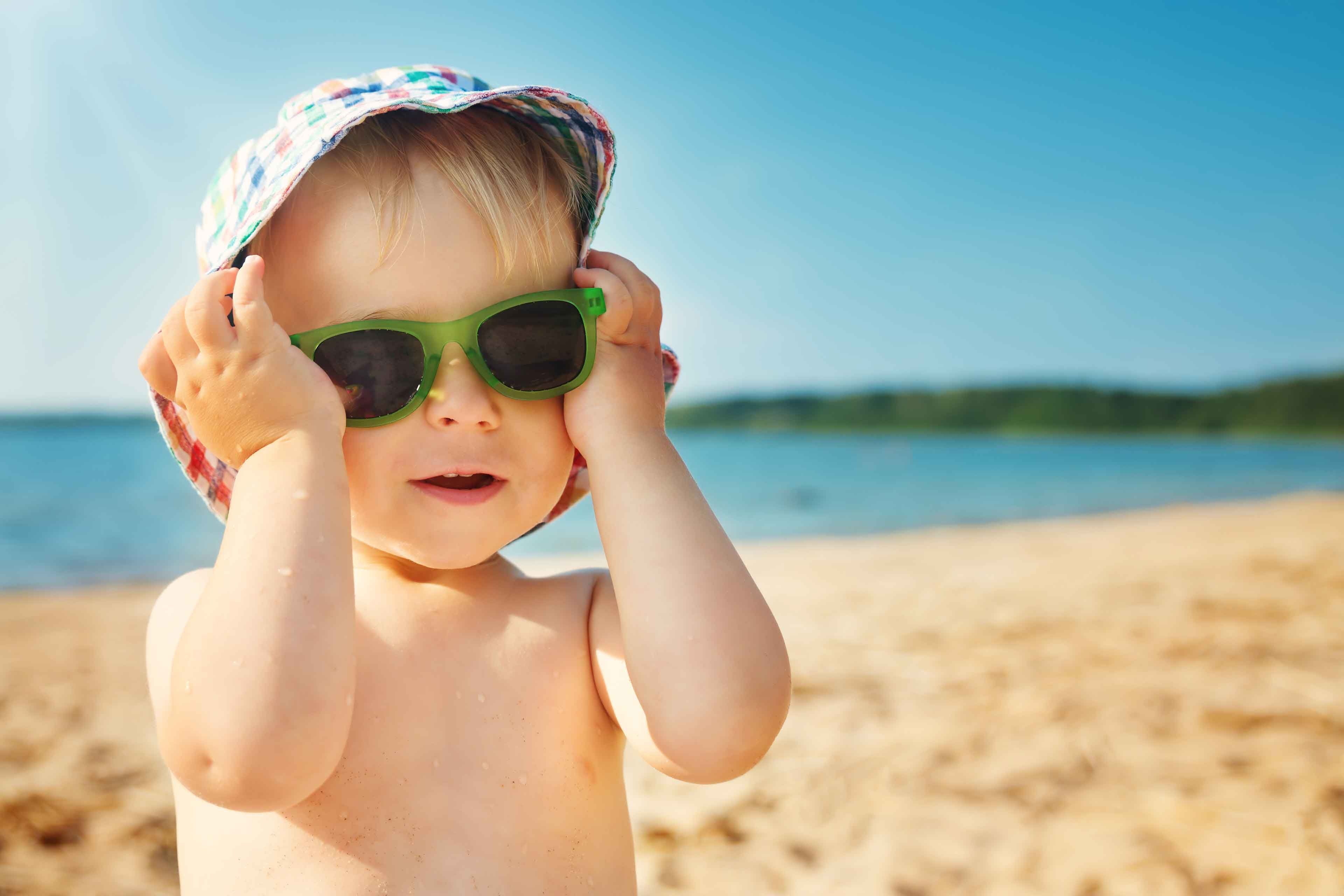 Ein kleines Kind am Strand, mit Sonnenbrille und Hut, mit einem unscharfen Hintergrund aus Meer und Sand.