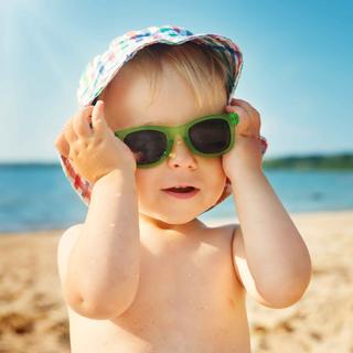 Ein kleines Kind am Strand, mit Sonnenbrille und Hut, mit einem unscharfen Hintergrund aus Meer und Sand.