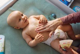 Lächelndes Baby auf der Wickelauflage, dem ein Elternteil sanft HiPP Öl-Balsam auf den Bauch einmassiert.