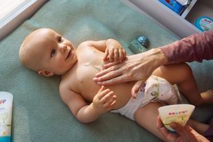 Lächelndes Baby auf der Wickelauflage, dem ein Elternteil sanft HiPP Öl-Balsam auf den Bauch einmassiert.