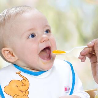 Ein Baby mit blauen Augen, das ein Lätzchen mit einem Elefantenmuster trägt und von einem Erwachsenen mit einem Löffel gefüttert wird.