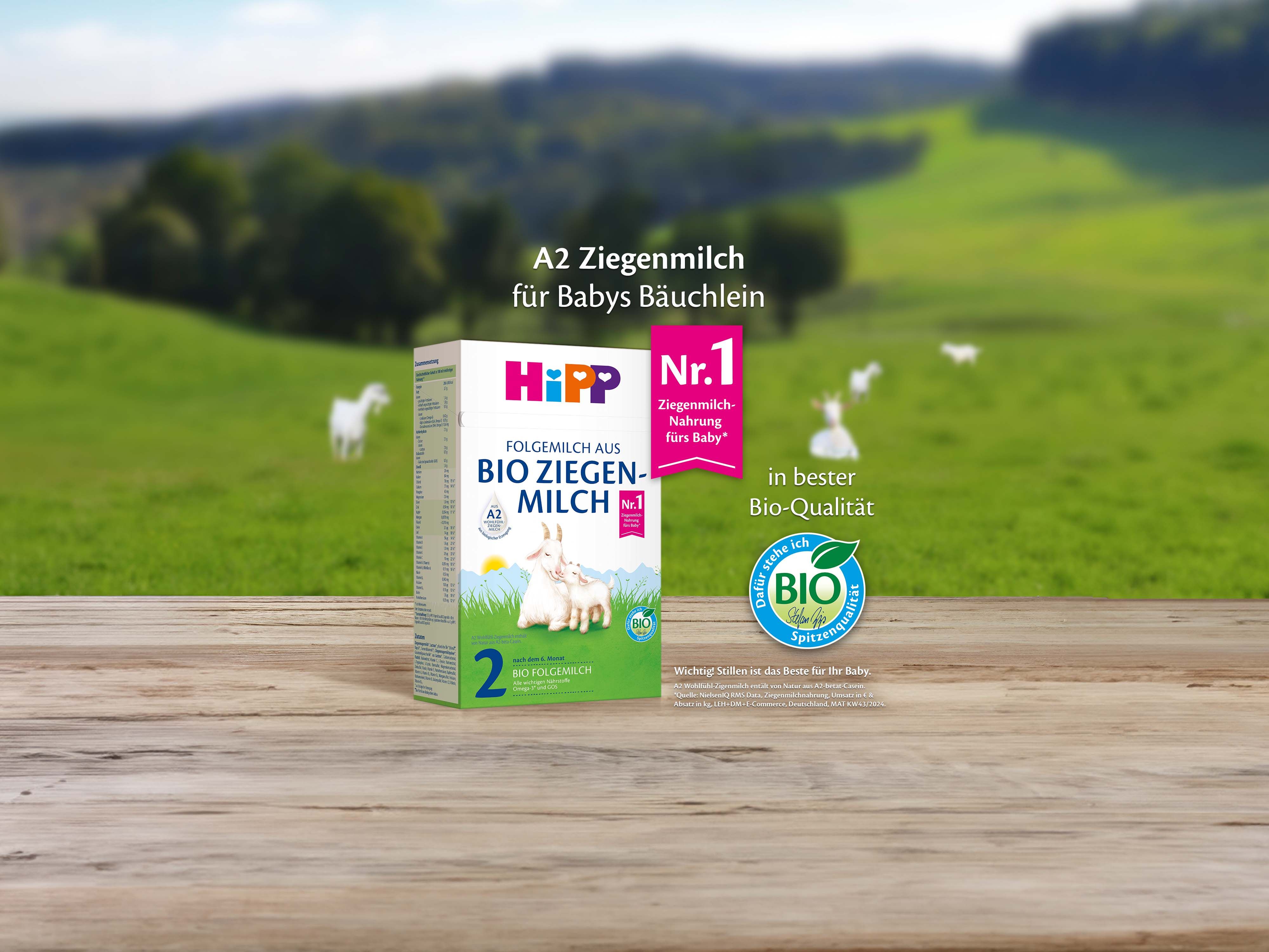 HiPP 2 Folgemilch aus Bio Ziegenmilch Packung auf Holztisch vor grüner Weidelandschaft mit Ziegenmotiv