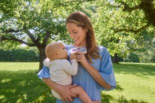 Mutter füttert ihr Baby im Grünen mit einer Flasche Milchnahrung von HiPP und lächelt es liebevoll an