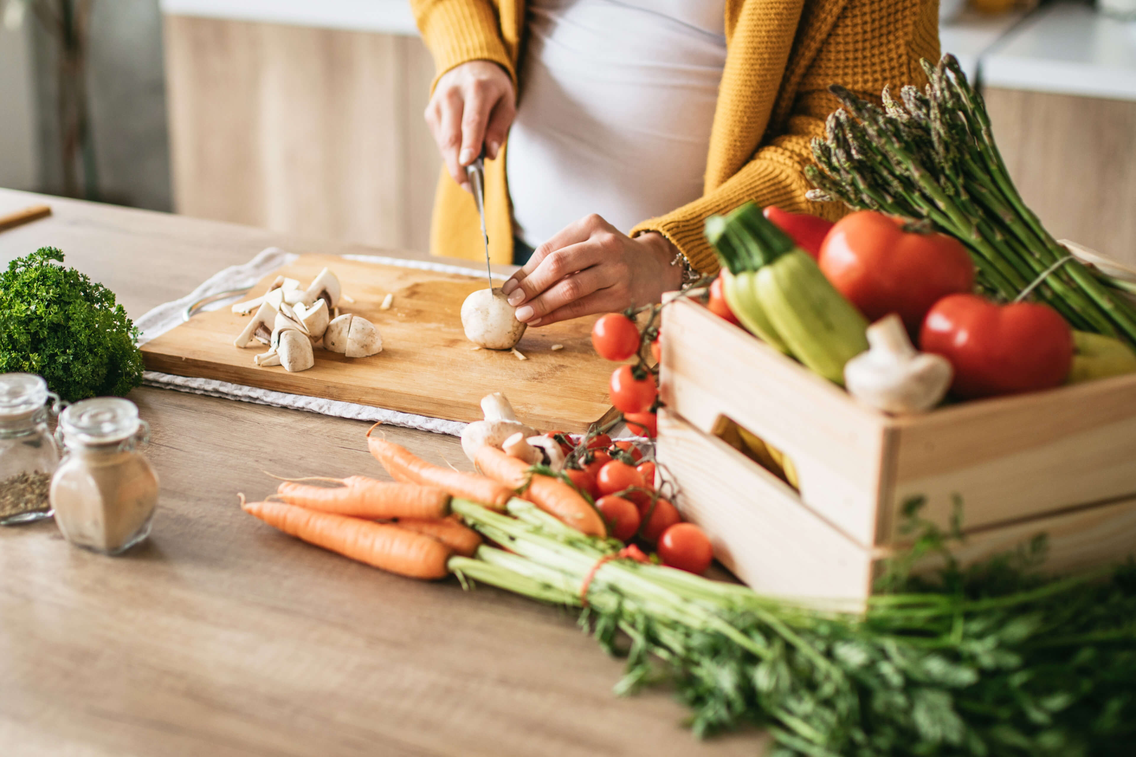 Eine Holzkiste mit frischem Gemüse, darunter Tomaten, Spargel und Knoblauch, auf einem Küchentisch mit verschiedenen Gewürzen und Utensilien im Hintergrund.
