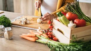 Eine Holzkiste mit frischem Gemüse, darunter Tomaten, Spargel und Knoblauch, auf einem Küchentisch mit verschiedenen Gewürzen und Utensilien im Hintergrund.