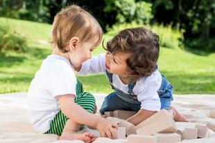 Zwei kleine Kinder, eines in einem weißen Hemd und das andere in einem blauen Overall, spielen im Freien auf einer Decke, wobei das Kind im blauen Overall den Fuß des anderen Kindes berührt.
