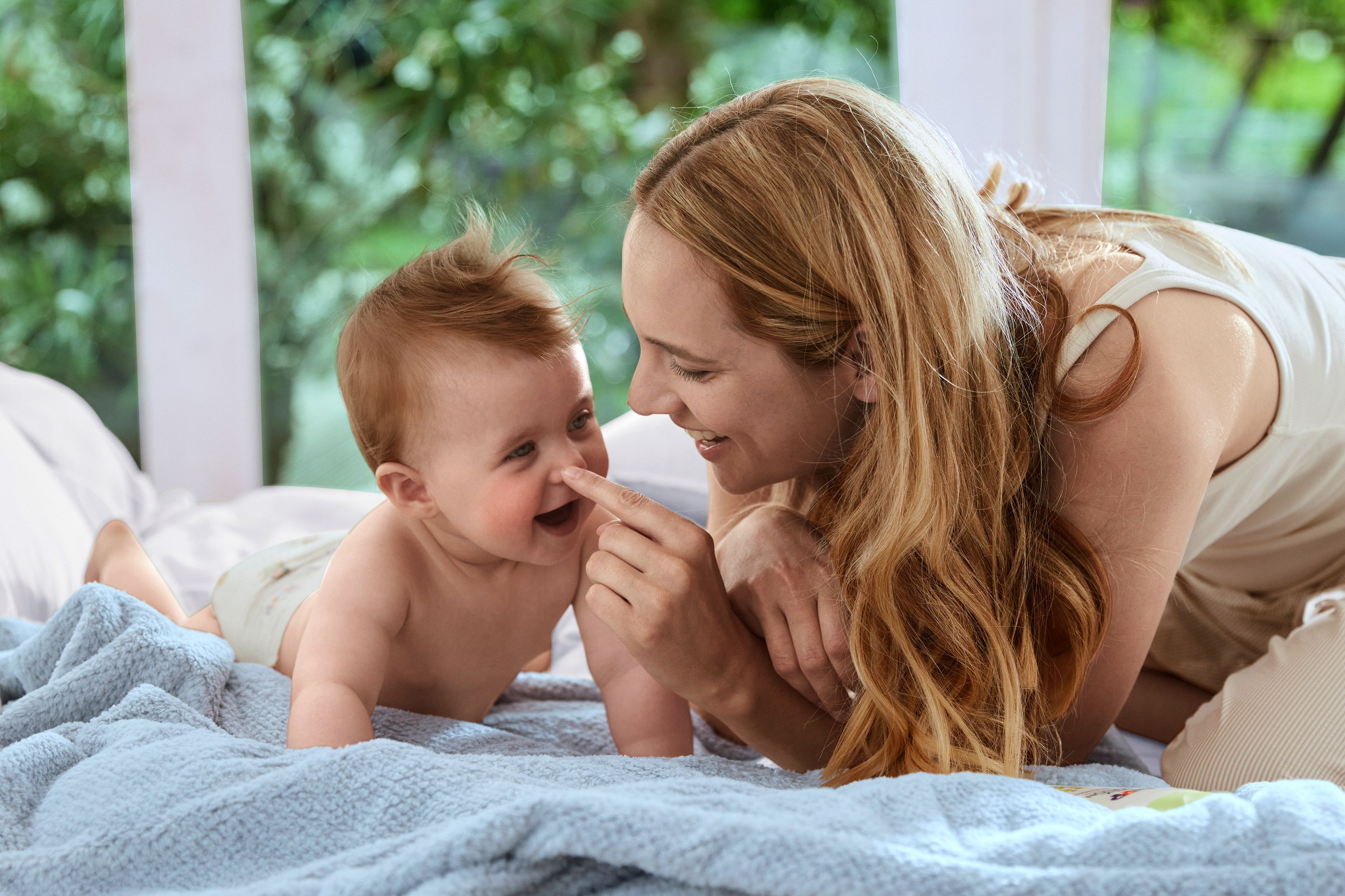 Ein zärtlicher Moment zwischen einer Mutter und ihrem Baby, in dem die Mutter spielerisch mit dem Baby auf einem Bettchen interagiert.