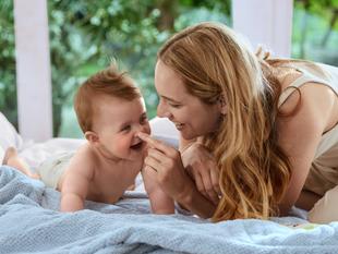 Ein zärtlicher Moment zwischen einer Mutter und ihrem Baby, in dem die Mutter spielerisch mit dem Baby auf einem Bettchen interagiert.