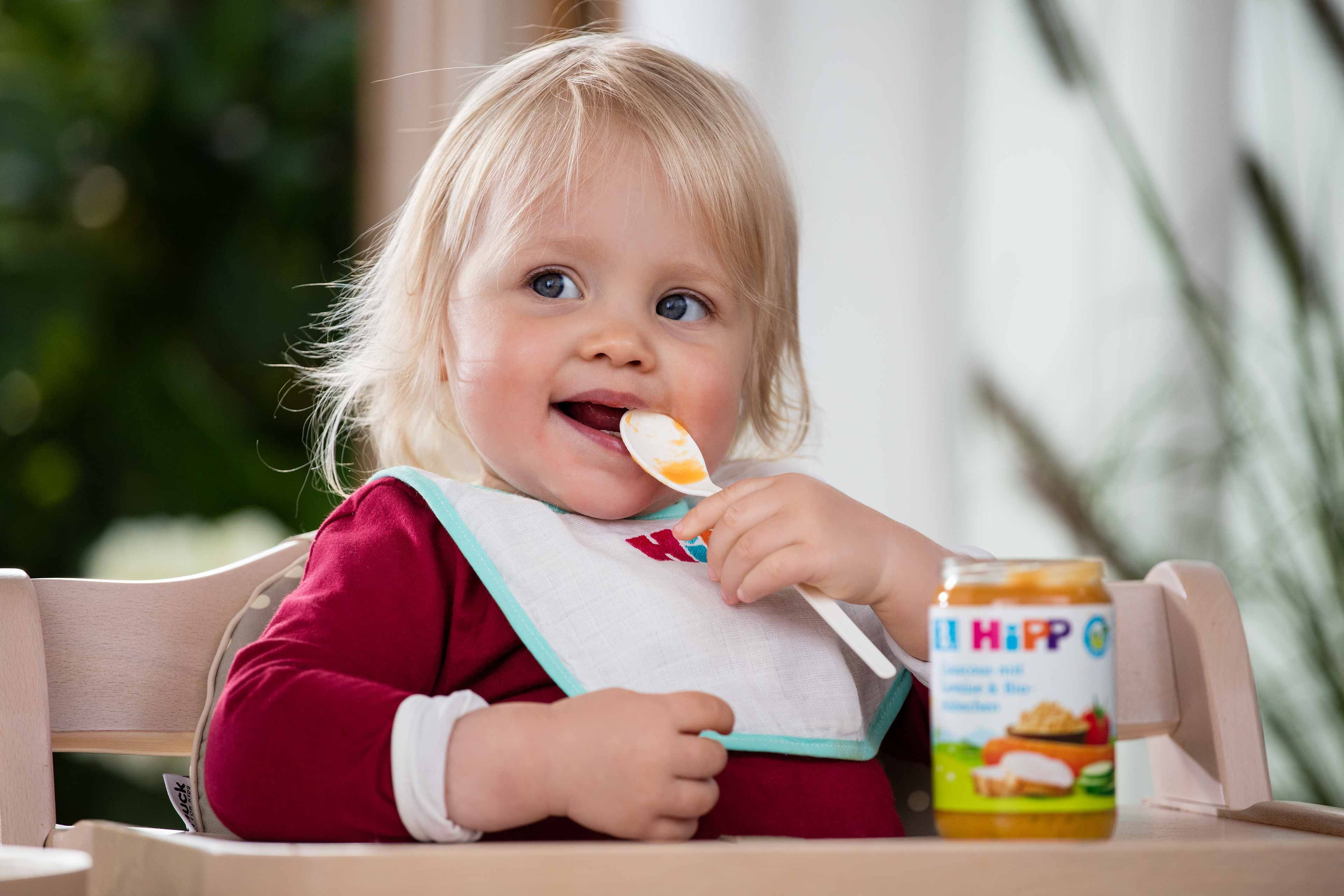 Ein kleines Kind mit blondem Haar, das ein Lätzchen trägt, einen Löffel hält und zu essen scheint.
