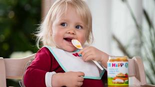 Ein kleines Kind mit blondem Haar, das ein Lätzchen trägt, einen Löffel hält und zu essen scheint.