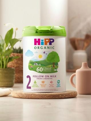 HiPP Organic Follow on Milk tub on a bright kitchen worktop beside a baby cup, dummy and plant, suggesting everyday family use