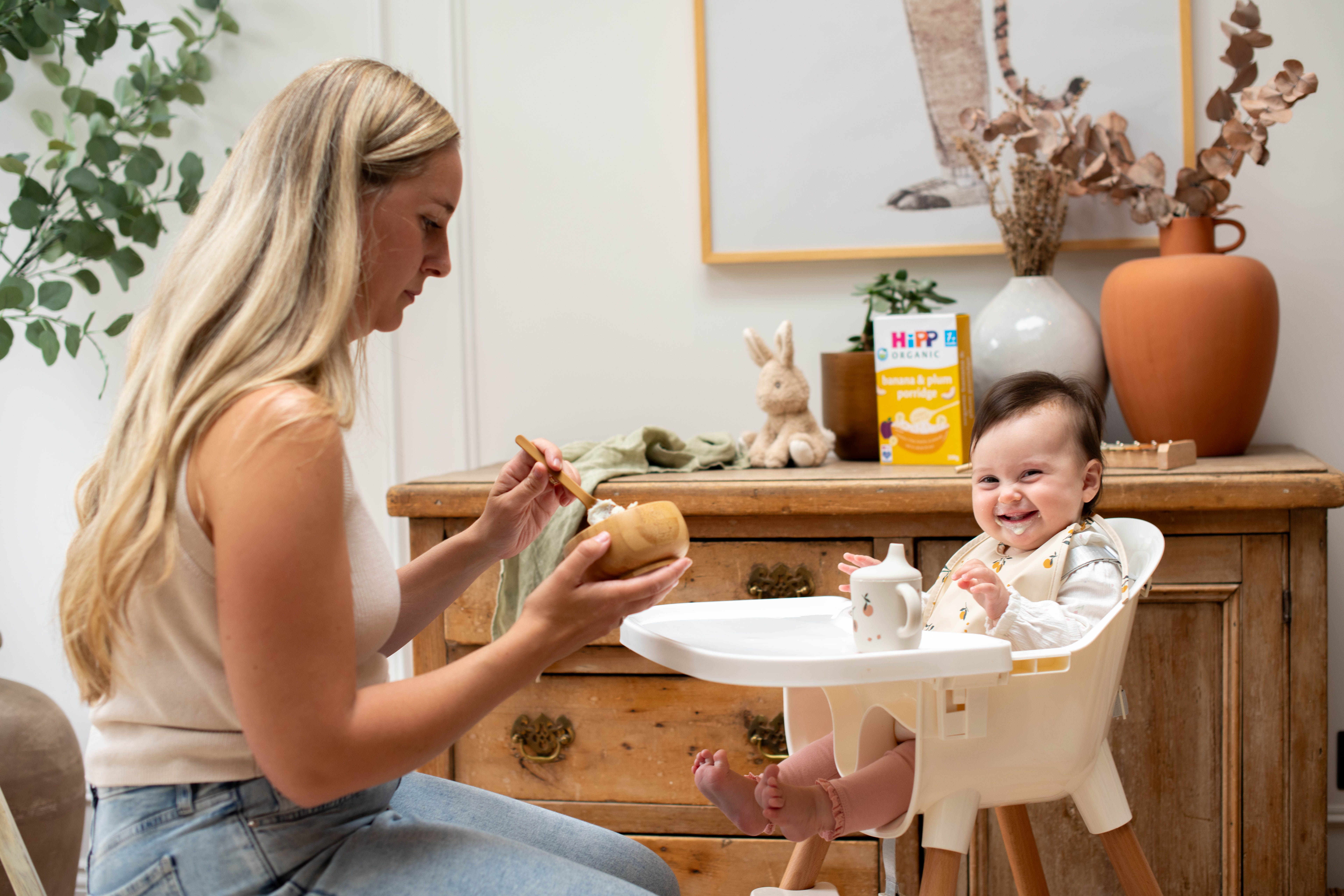 Smiling baby in a highchair being spoon-fed by mum, with HiPP Organic baby food on a wooden sideboard behind them