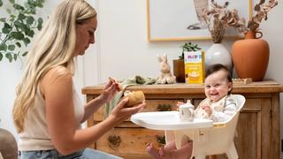 Smiling baby in a highchair being spoon-fed by mum, with HiPP Organic baby food on a wooden sideboard behind them