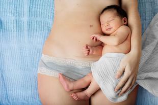 Eine Mutter hält ihr neugeborenes Baby, das in ein weißes Tuch gewickelt ist, während die Mutter hellblaue Unterwäsche mit Spitzenverzierung trägt.