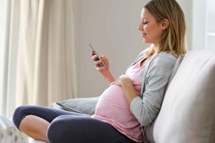 Schwangere Frau sitzt entspannt auf dem Sofa, hält ihre Hand auf den Bauch und liest lächelnd auf ihrem Smartphone.
