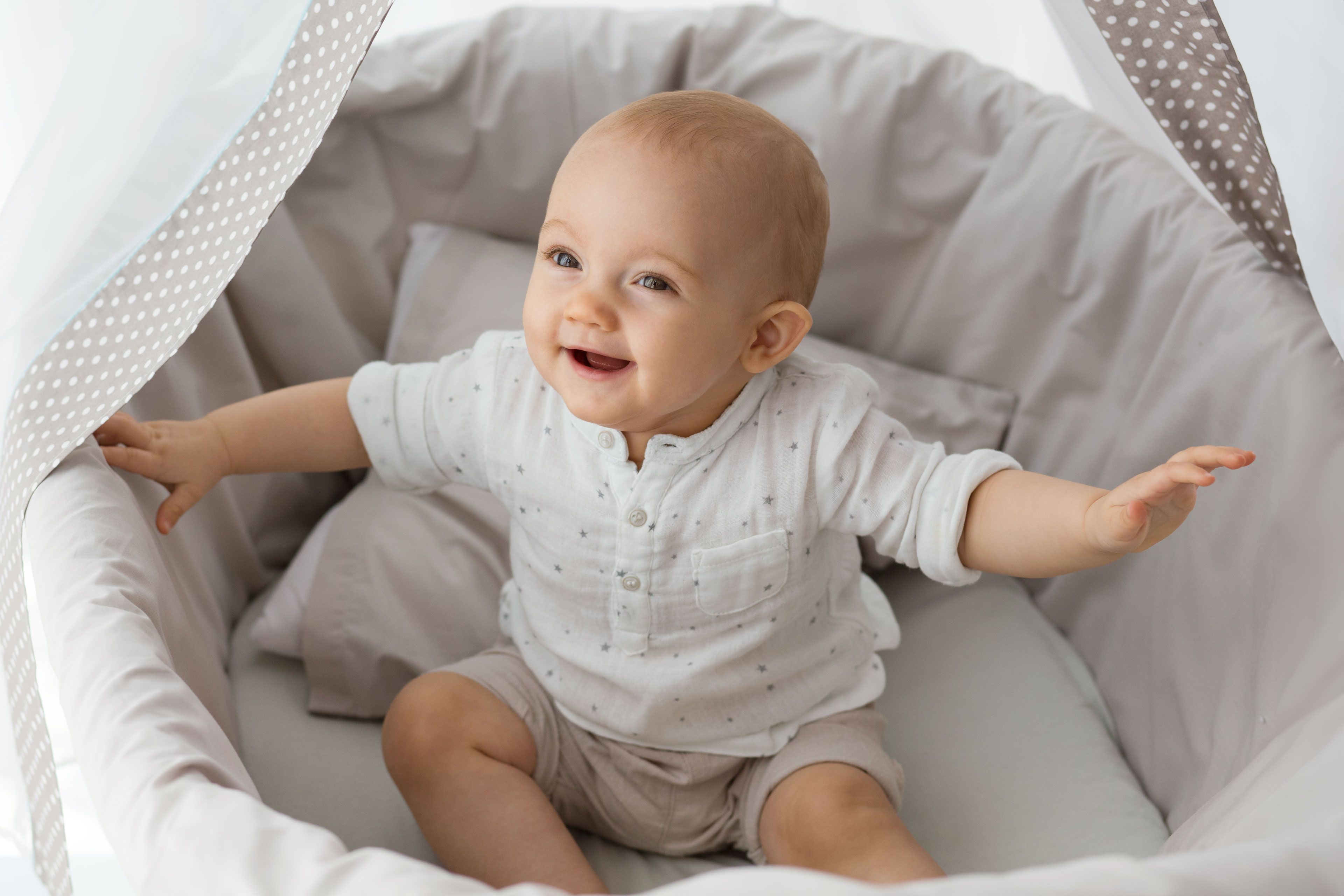Ein weiß gekleidetes Baby sitzt in einem gepunkteten Bettchen und schaut neugierig zur Seite.