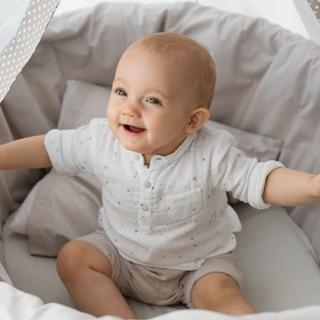 Ein weiß gekleidetes Baby sitzt in einem gepunkteten Bettchen und schaut neugierig zur Seite.