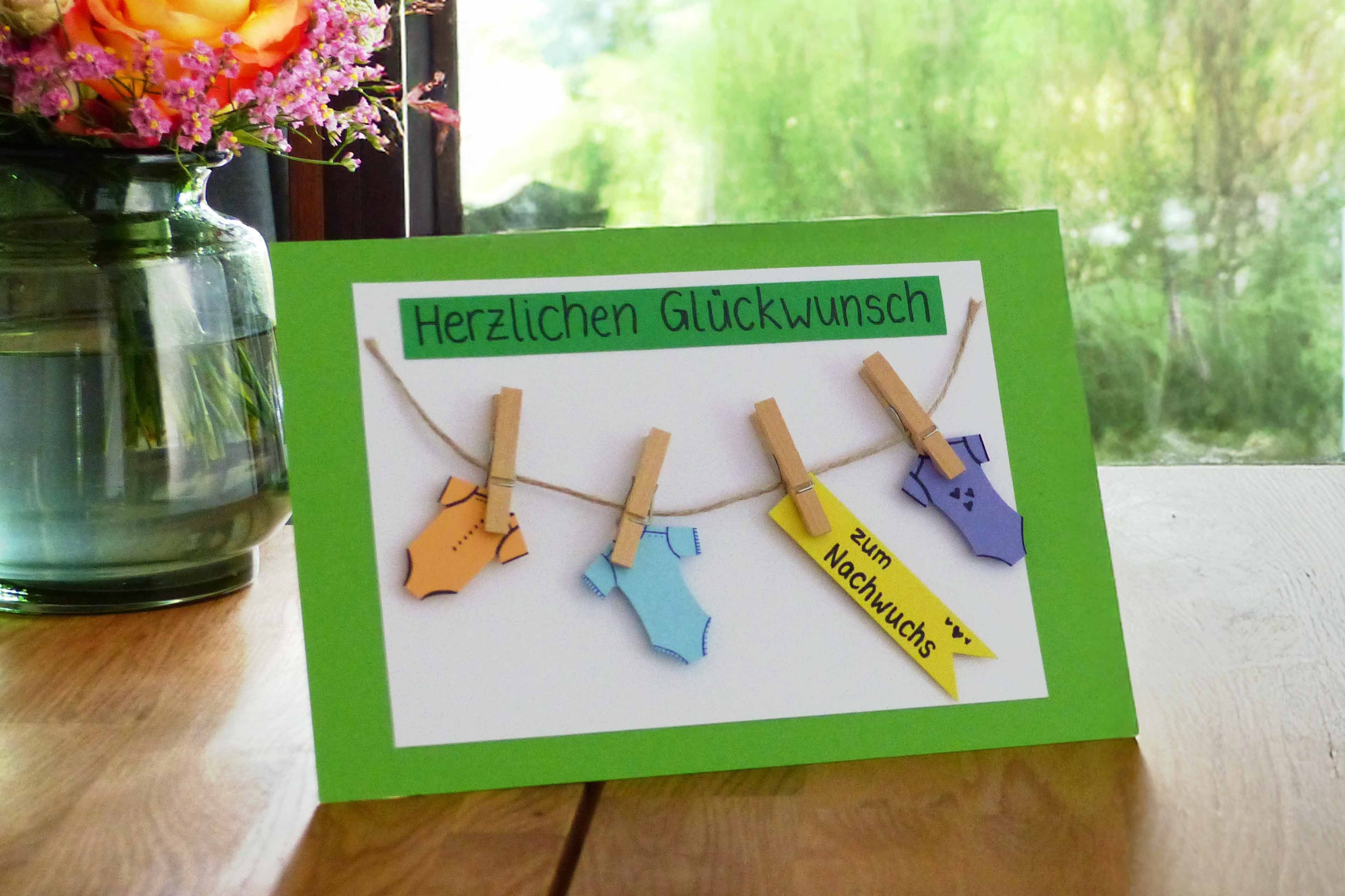 Eine handgefertigte Karte mit dem deutschen Text "Herzlichen Glückwunsch" am oberen Rand. Unter dem Text hängen vier Baby-Strampler an einer Wäscheleine, die jeweils mit einem Namensschild versehen sind. Die Karte ist vor einem grünen Hintergrund auf einer Holzunterlage platziert.