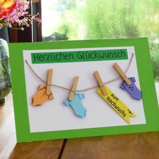 Eine handgefertigte Karte mit dem deutschen Text "Herzlichen Glückwunsch" am oberen Rand. Unter dem Text hängen vier Baby-Strampler an einer Wäscheleine, die jeweils mit einem Namensschild versehen sind. Die Karte ist vor einem grünen Hintergrund auf einer Holzunterlage platziert.