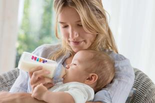 Frau hält ein Baby im Arm und füttert es mit einer Flasche mit dem Aufdruck 'HiPP'.