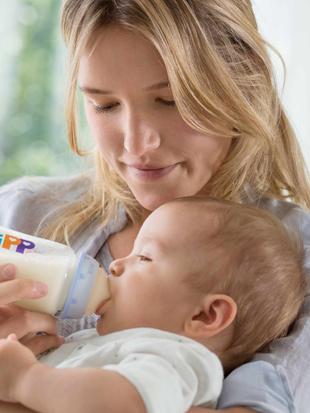 Frau hält ein Baby im Arm und füttert es mit einer Flasche mit dem Aufdruck 'HiPP'.