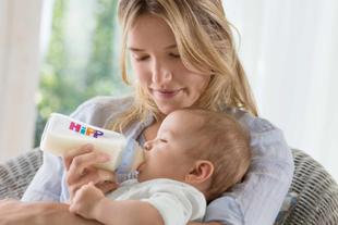 Frau hält ein Baby im Arm und füttert es mit einer Flasche mit dem Aufdruck 'HiPP'.