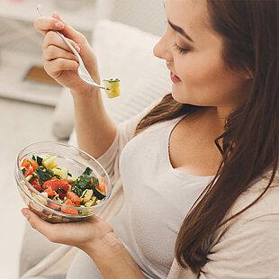 A woman sits comfortably indoors, smiling as she eats a fresh salad from a glass bowl. The salad includes colorful vegetables like tomatoes, cucumbers, spinach, and possibly pasta.