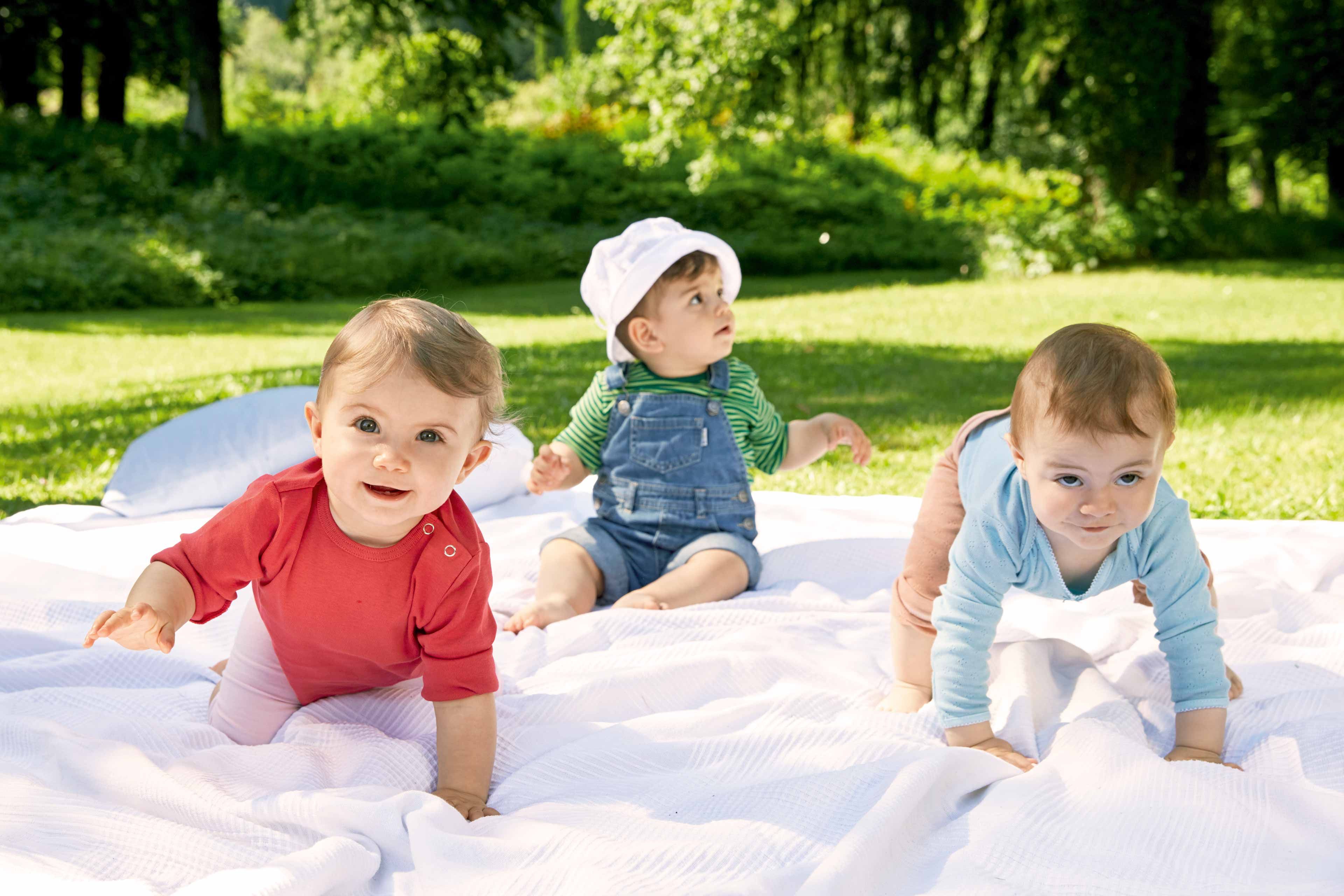 Drei Babys im Freien auf einer Decke, eines mit rotem Hemd, ein anderes in einem blauen Overall und das dritte mit einer weißen Mütze.