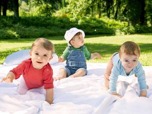 Drei Babys im Freien auf einer Decke, eines mit rotem Hemd, ein anderes in einem blauen Overall und das dritte mit einer weißen Mütze.