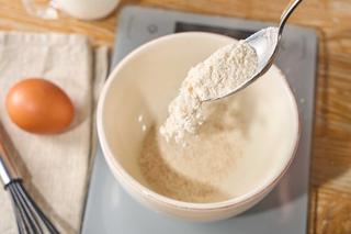 Ein Löffel lässt helles Mehl in eine beige Schüssel rieseln, die auf einer Küchenwaage steht, daneben liegen ein braunes Ei und ein Schneebesen.