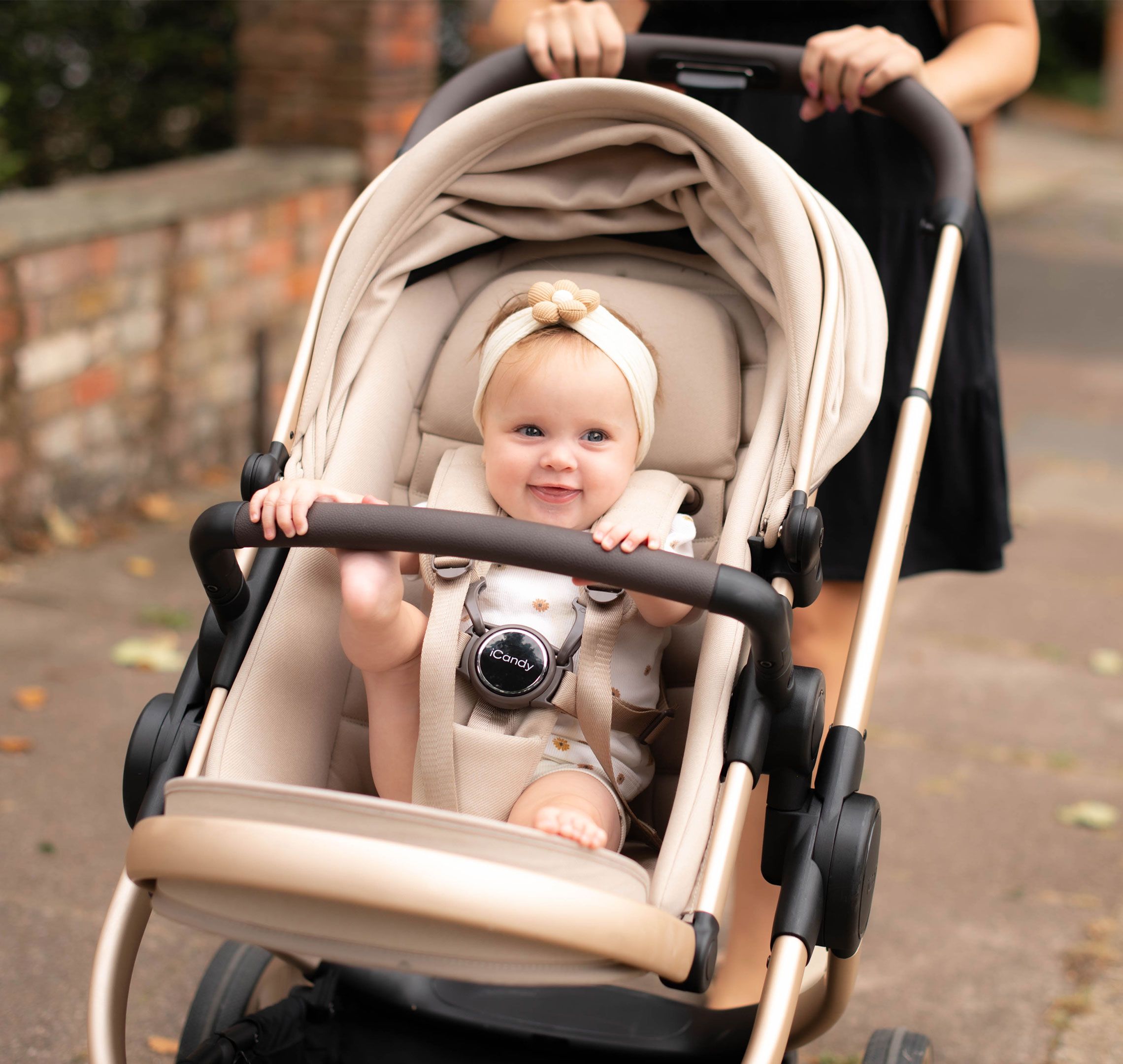 A smiling baby sitting in a beige pram outdoors, wearing a cream headband with a flower detail and secured in the stroller harness, while an adult pushes the pram along a paved path in a residential setting.