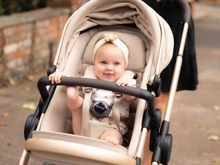 A smiling baby sitting in a beige pram outdoors, wearing a cream headband with a flower detail and secured in the stroller harness, while an adult pushes the pram along a paved path in a residential setting.