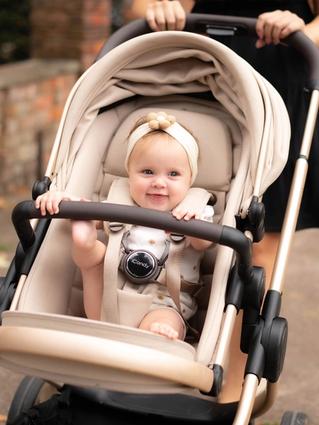 A smiling baby sitting in a beige pram outdoors, wearing a cream headband with a flower detail and secured in the stroller harness, while an adult pushes the pram along a paved path in a residential setting.
