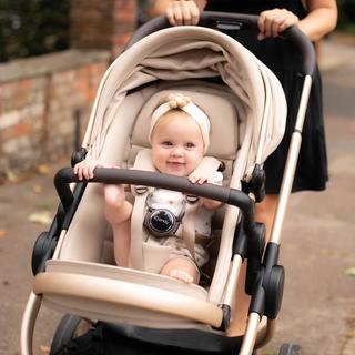 A smiling baby sitting in a beige pram outdoors, wearing a cream headband with a flower detail and secured in the stroller harness, while an adult pushes the pram along a paved path in a residential setting.