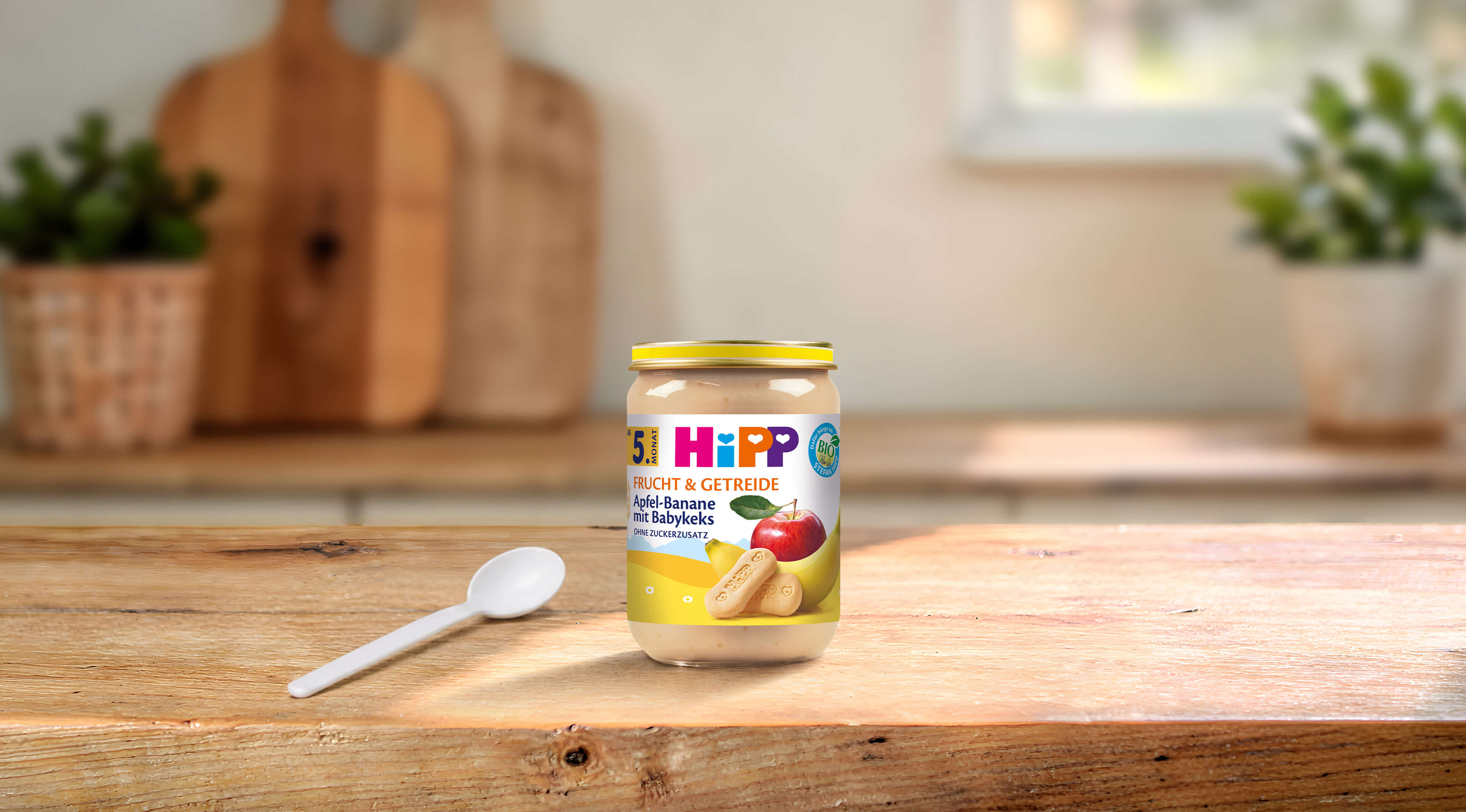 HiPP Bio-Gläschen Apfel-Banane mit Babykeks 190g auf Holztisch mit Löffel in heller, gemütlicher Küchenumgebung