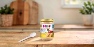 HiPP Bio-Gläschen Apfel-Banane mit Babykeks 190g auf Holztisch mit Löffel in heller, gemütlicher Küchenumgebung