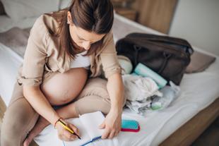 Eine schwangere Frau schreibt in ein Notizbuch, während sie auf einem Bett sitzt, umgeben von verschiedenen Gegenständen, darunter eine Tasche und eine Bürste.