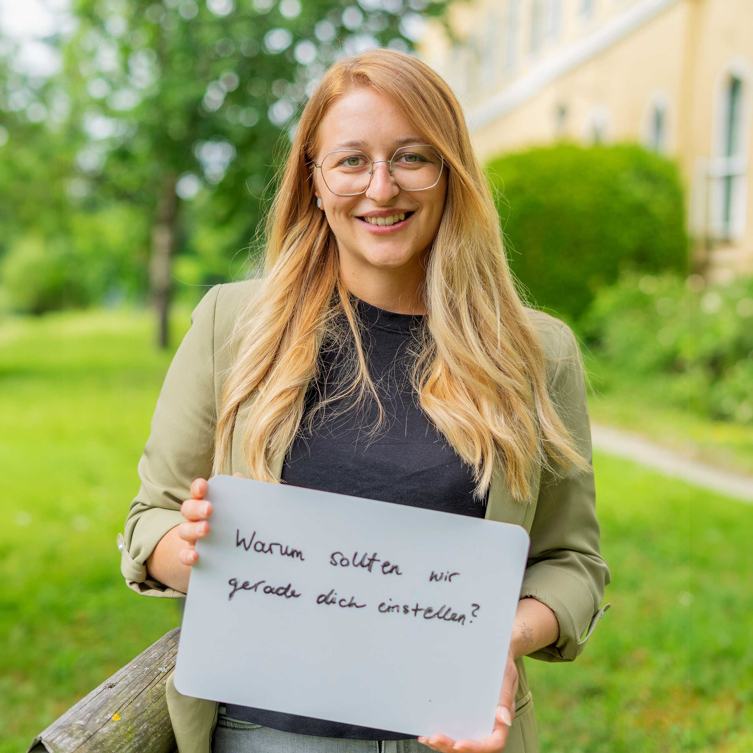 Eine HiPP Mitarbeiterin hält ein Schild mit 'Warum sollten wir gerade dich einstellen?'