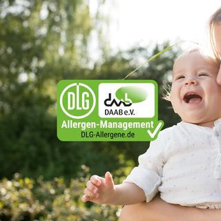 Lachendes Baby im Arm der Mutter im Grünen, daneben ein DLG-Allergen-Management-Siegel für sicheres Allergenmanagement.
