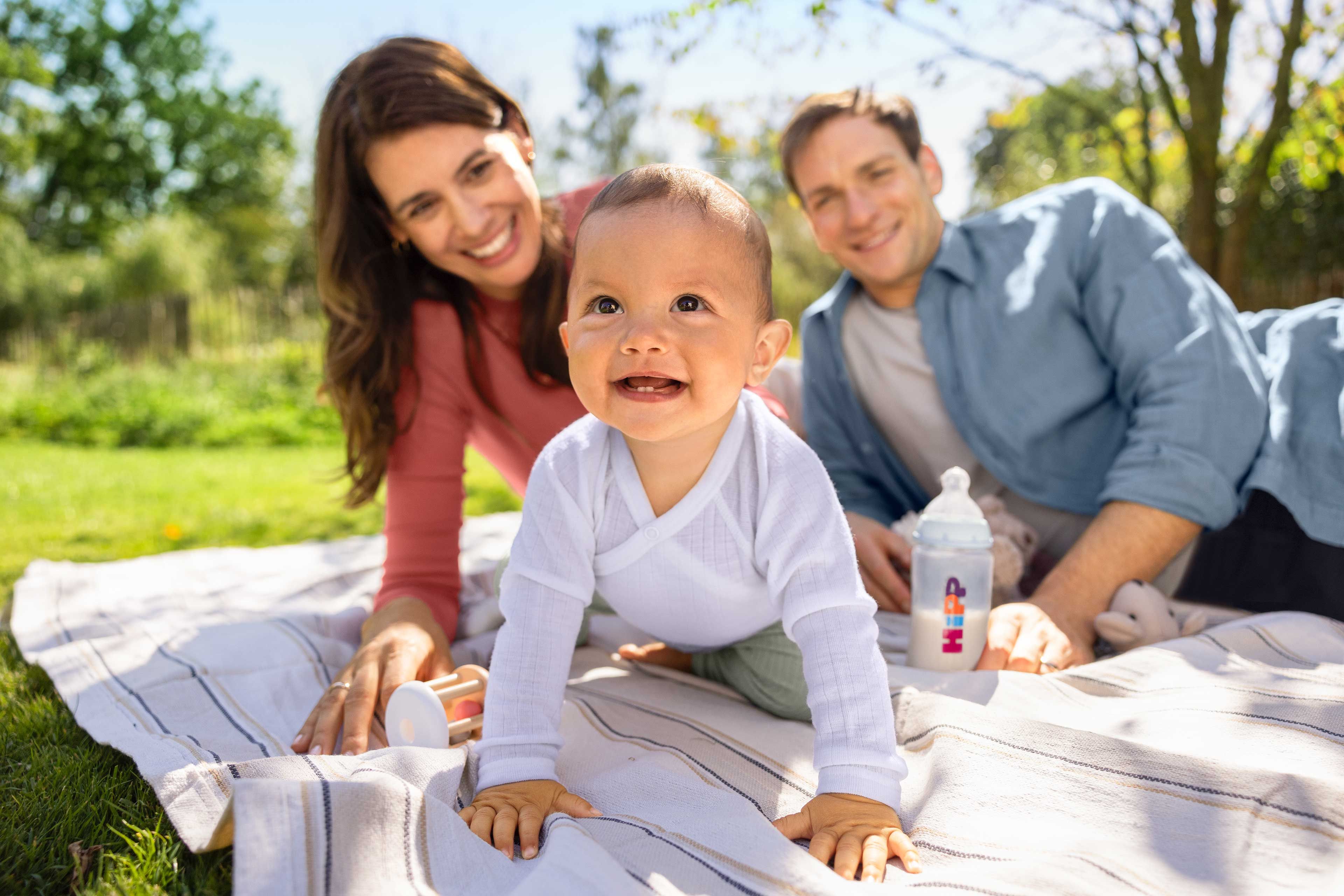 Lachendes Baby krabbelt auf einer Picknickdecke im Grünen, Eltern dahinter mit HiPP Milchnahrung in der Flasche.