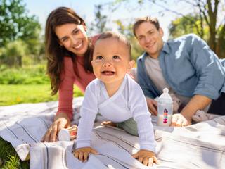 Lachendes Baby krabbelt auf einer Picknickdecke im Grünen, Eltern dahinter mit HiPP Milchnahrung in der Flasche.