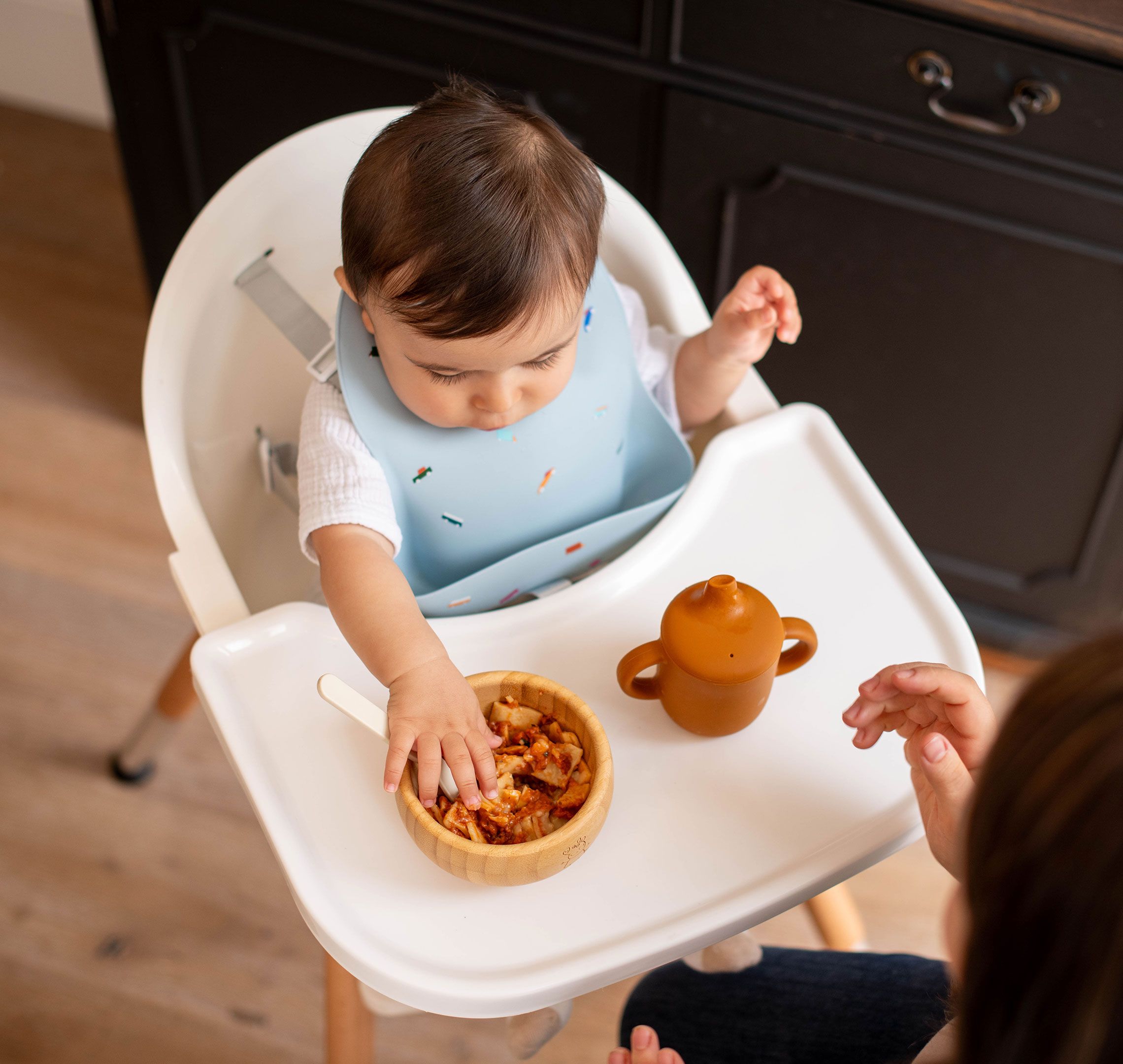 Baby in a highchair exploring a bowl of lumpy food with their hands during weaning, with a parent close by for support