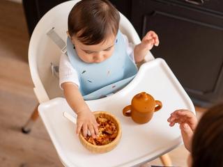 Baby in a highchair exploring a bowl of lumpy food with their hands during weaning, with a parent close by for support