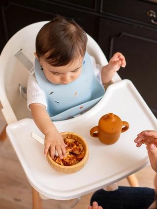 Baby in a highchair exploring a bowl of lumpy food with their hands during weaning, with a parent close by for support