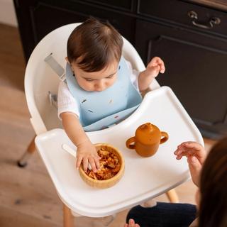 Baby in a highchair exploring a bowl of lumpy food with their hands during weaning, with a parent close by for support
