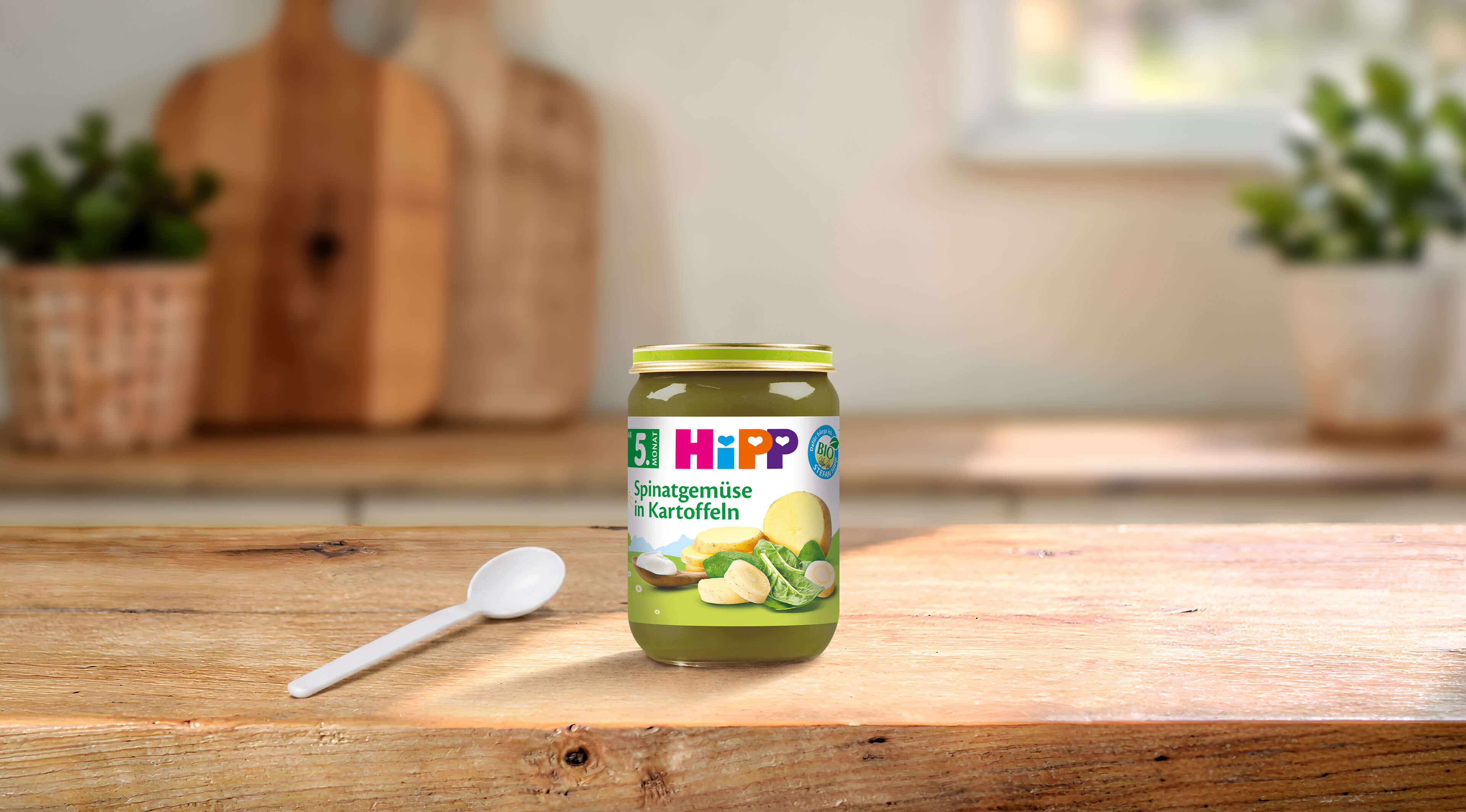 HiPP Bio-Gläschen „Spinatgemüse in Kartoffeln“ ab 5. Monat auf Holztisch, daneben ein Löffel in heller Küchenumgebung