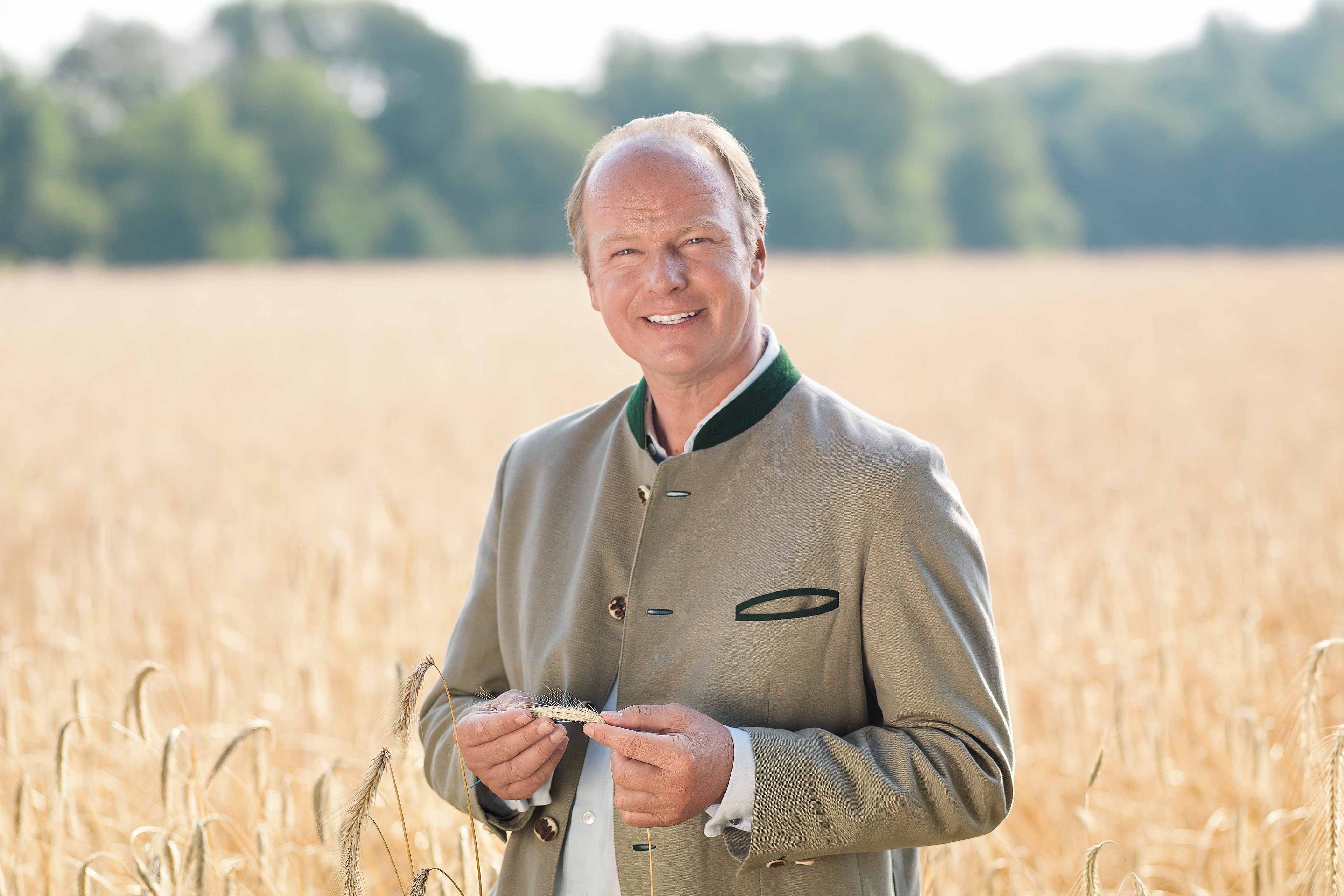 Lächelnder Mann im Trachtenjanker steht in einem goldenen Bio-Getreidefeld und hält Ähren in den Händen.
