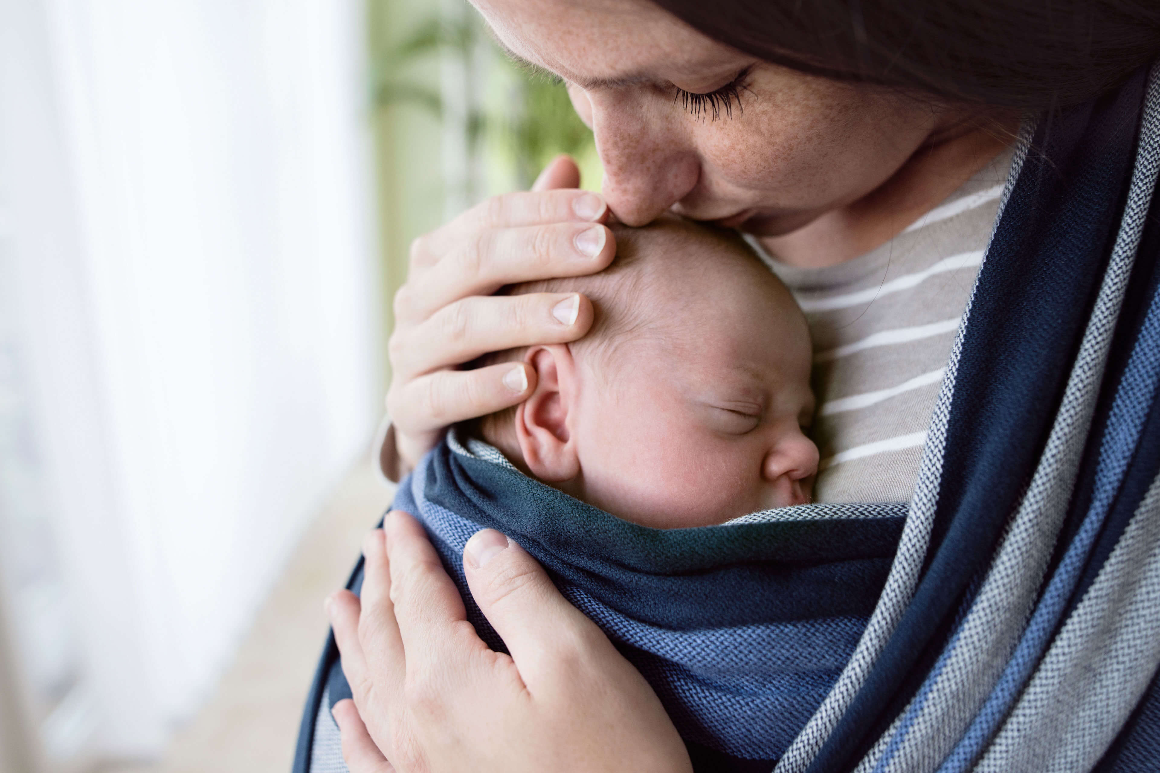 Nahaufnahme des Gesichts eines Babys, das in ein blaues Tuch gebettet ist und von der Hand eines Erwachsenen gehalten wird.